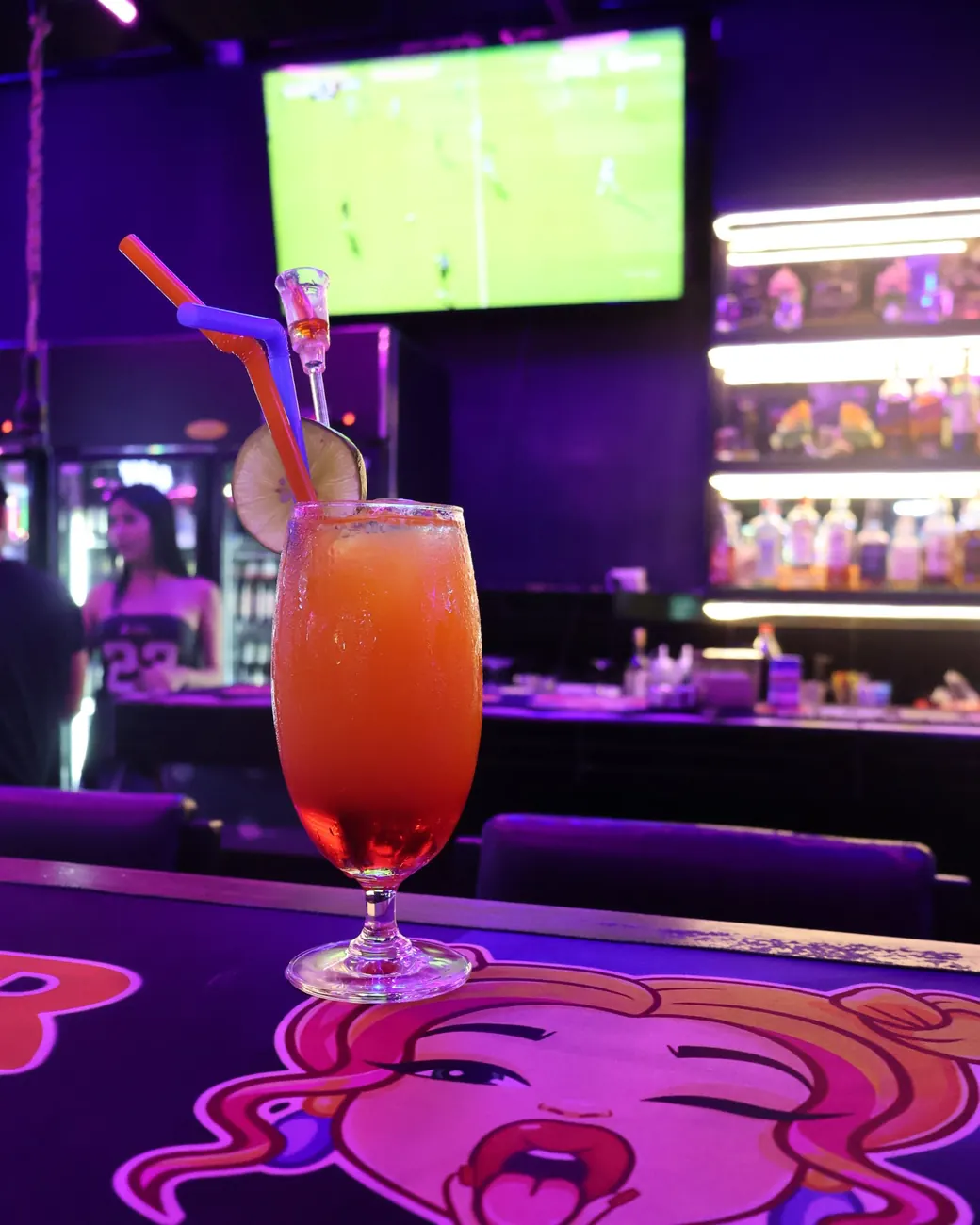 An orange and red cocktail on the bar, with football on the TV behind